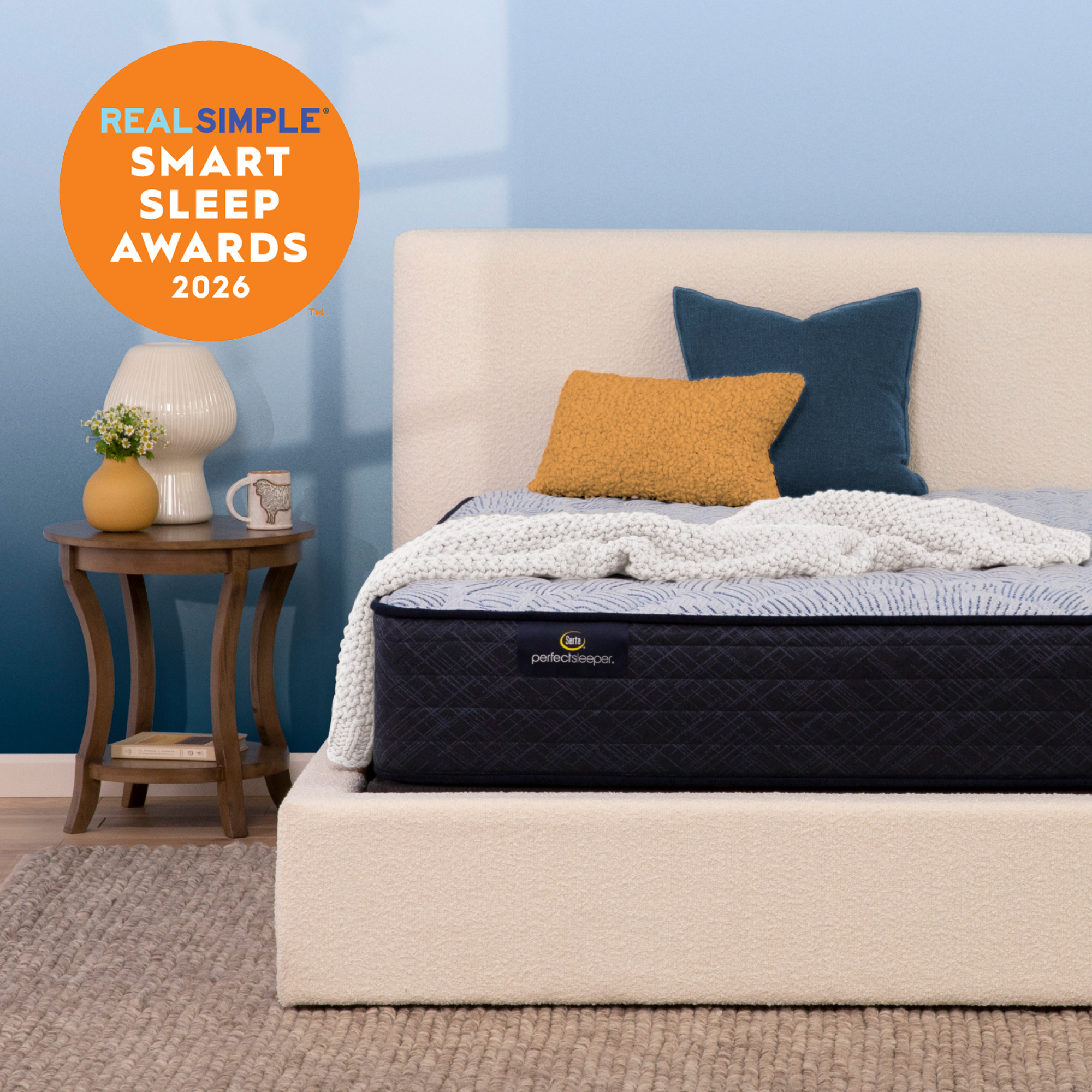 A Serta Perfect Sleeper firm mattress with a dark quilted, white textured bedding, and two pillows (one mustard yellow, one navy blue) in a styled bedroom setting with a blue wall, beige headboard, and a wooden side table. An orange badge in the top left reads 'REAL SIMPLE SMART SLEEP AWARDS 2026'.
