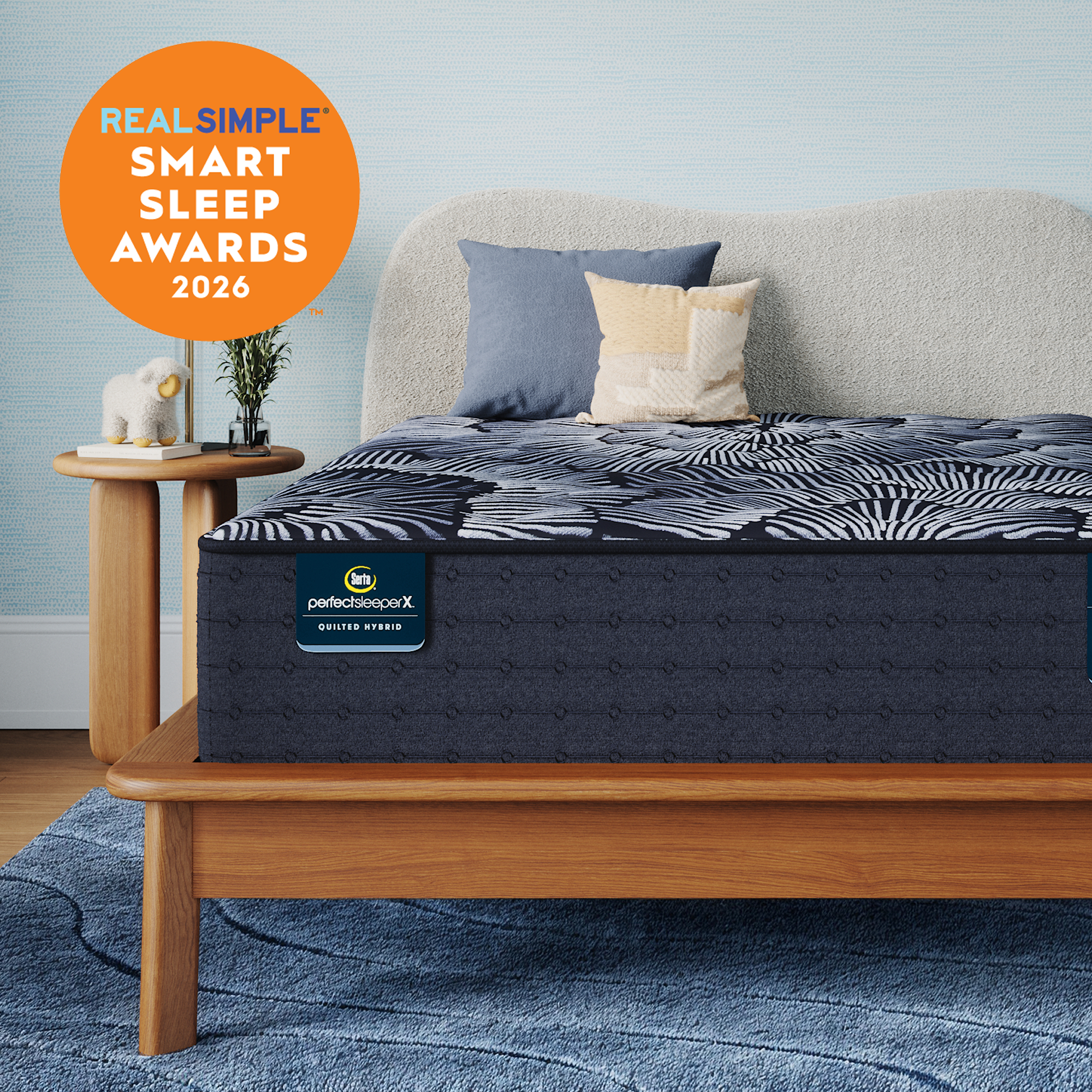 A Perfect Sleeper X Quilted Hybrid Excelled Plush mattress with a dark pattern and matching pillow shams styled in a bedroom setting, with a light wood bed frame, blue rug, side table, and a large orange badge reading 'REAL SIMPLE SMART SLEEP AWARDS 2026'. The mattress label shows 'perfectscore X' and 'BETTER START'.